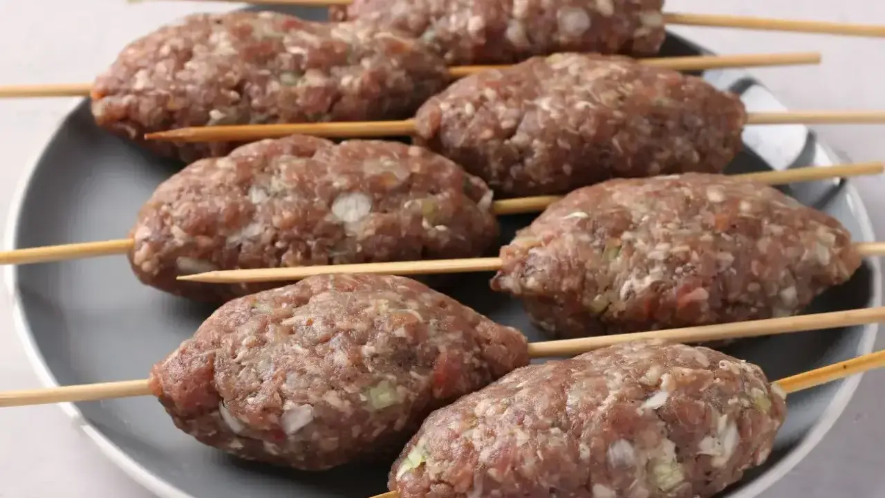Siedem surowych kofta kebab na patyczkach, ułożonych na szarym talerzu, gotowych do grillowania.