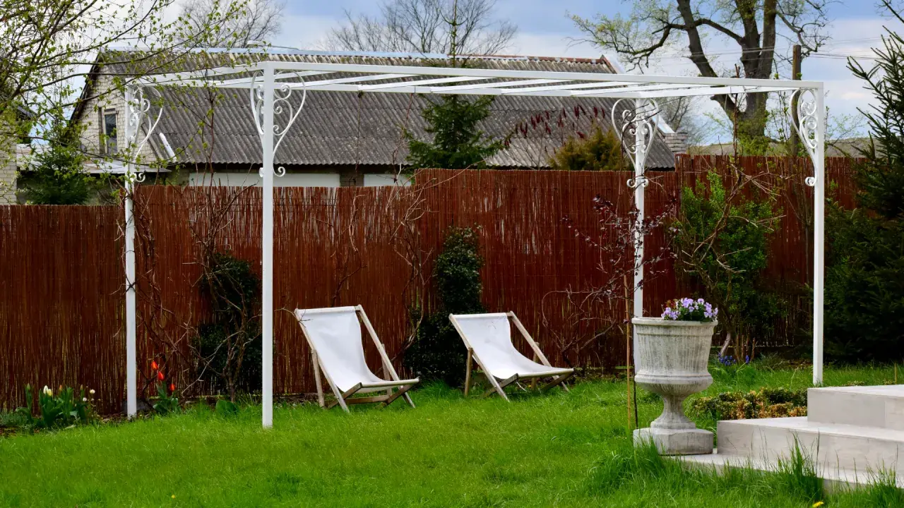 Biała pergola ogrodowa z dwoma leżakami i donicą z kwiatami.
