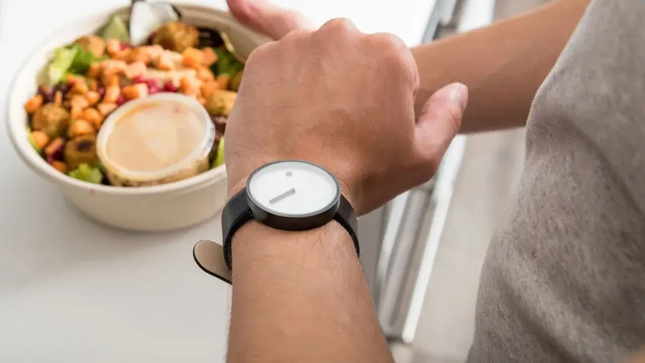 Mann schaut auf seine Uhr, neben einer Schüssel mit Salat. Die Frage: darf man fasten wenn man krank ist?