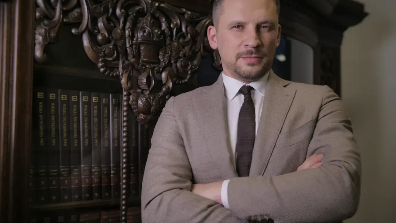 Mężczyzna w garniturze stoi przed regałem z książkami.