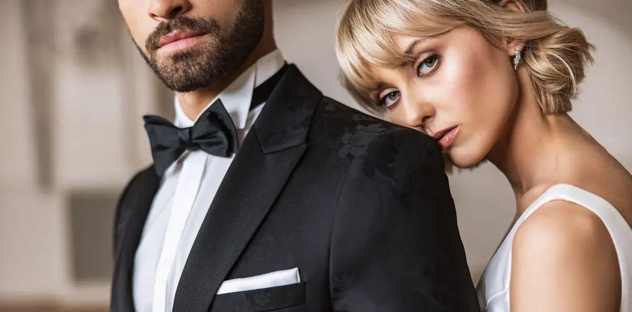 Elegancki mężczyzna w czarnym smokingu z muchą i białą koszulą. Obok niego opiera się kobieta w białej sukni. Idealny przykład, jaka koszula do muchy.
