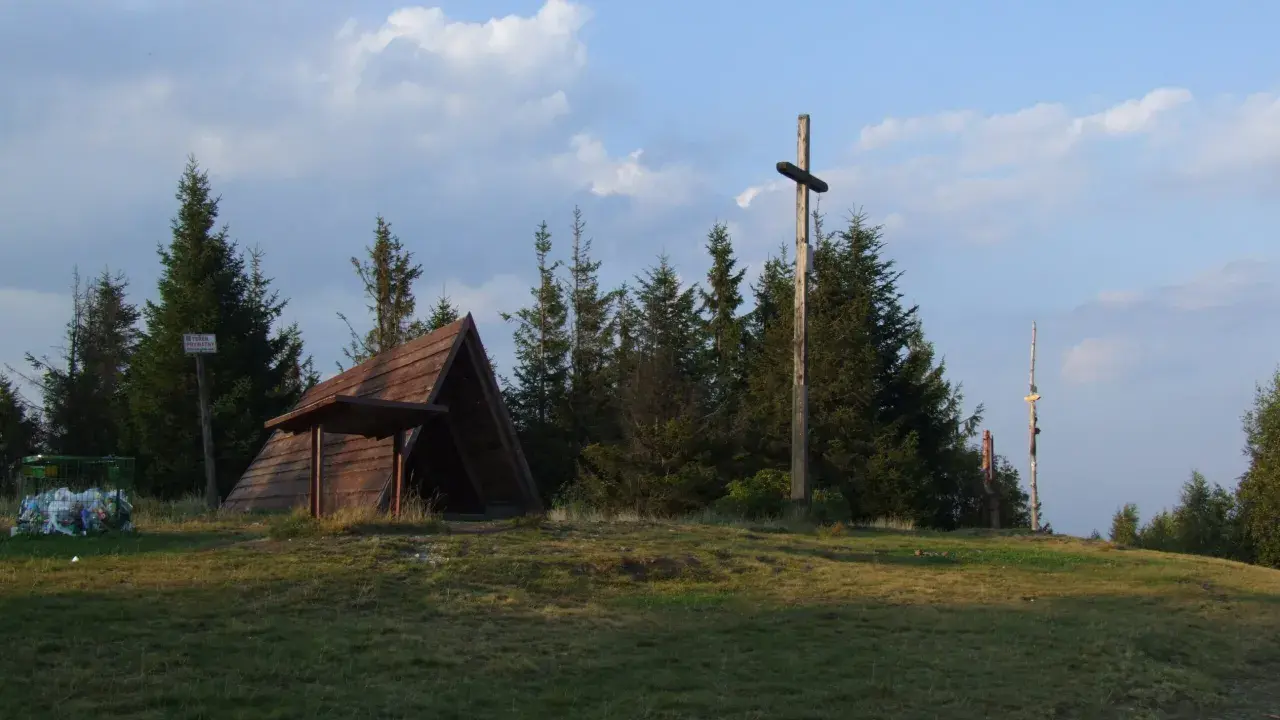 Drewniana wiata, krzyż i kosz na śmieci na tle drzew i nieba.