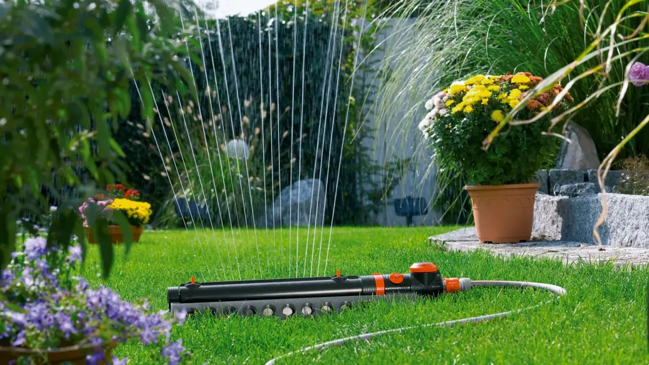 Sistema de riego automático Gardena en césped verde, con flores amarillas y moradas al fondo.