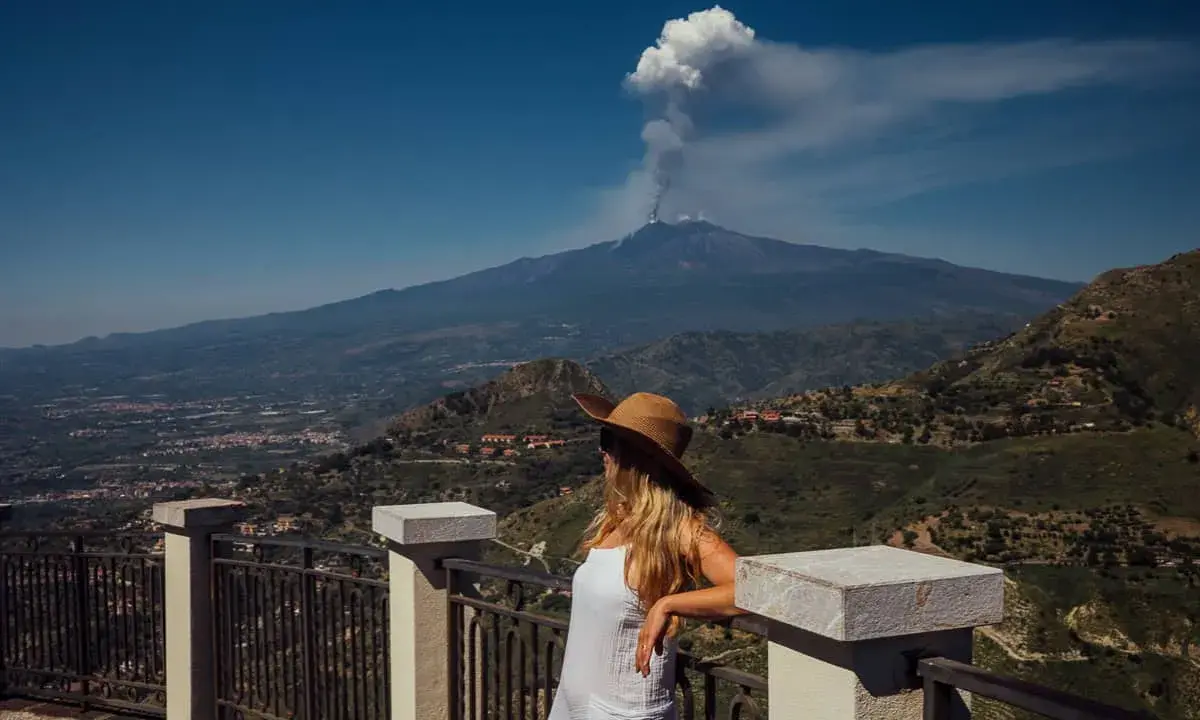 Frau mit Hut blickt auf den rauchenden sizilien vulkan. Die Landschaft ist grün und bergig.
