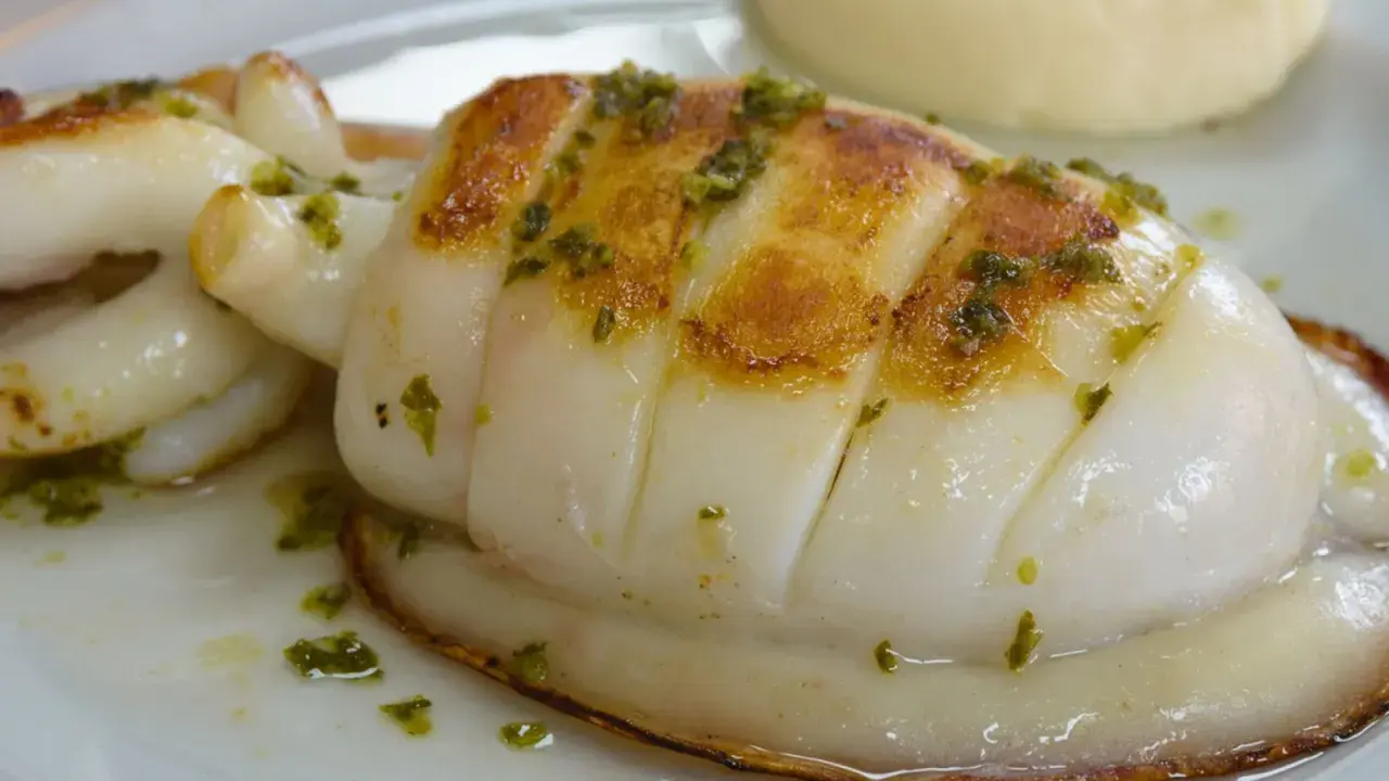 Delicioso sepia a la plancha con hierbas verdes, servido en un plato blanco con una guarnición cremosa.