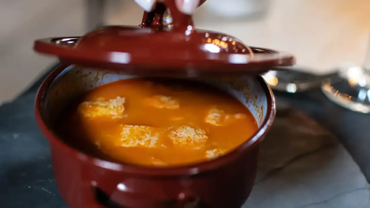 Una mano levanta la tapa de una cazuela de barro con una deliciosa comida típica de Ávila, un guiso naranja con trozos blancos.