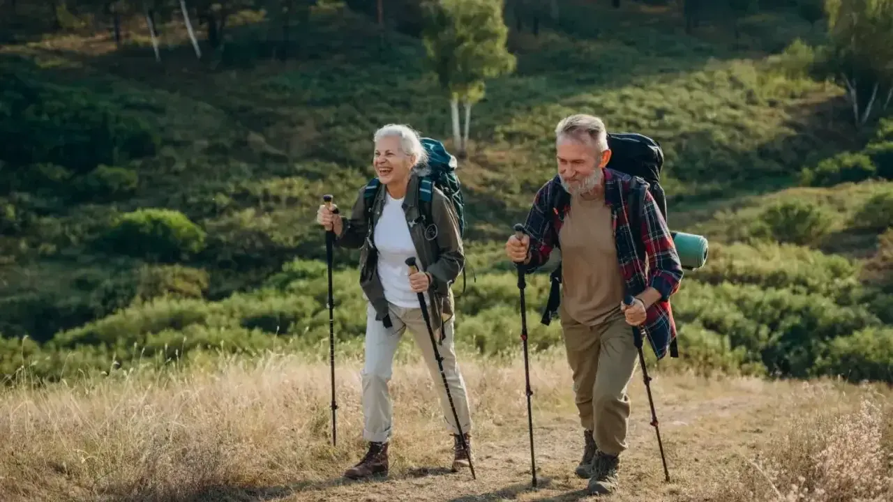 Uśmiechnięta para seniorów z plecakami przemierza leśny szlak, ucząc się, jak chodzić z kijkami.