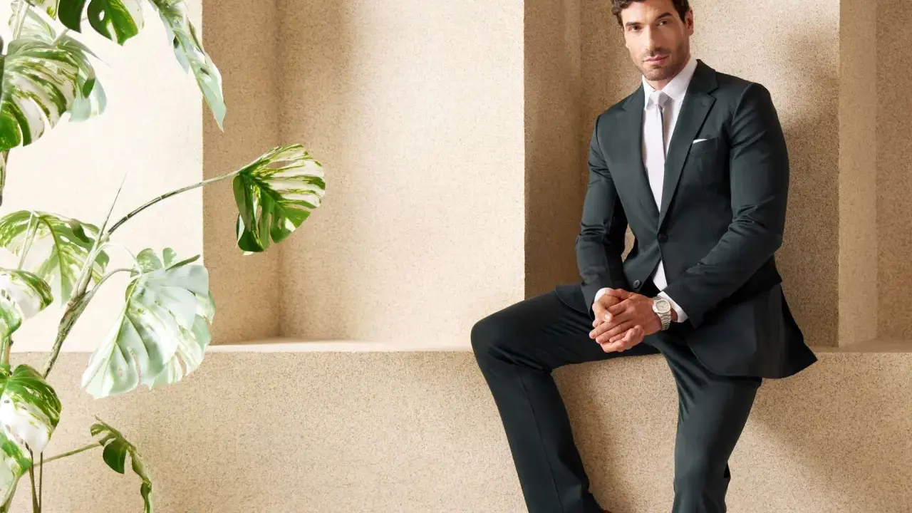 A man in a sharp green suit and black shirt poses confidently next to a lush plant.