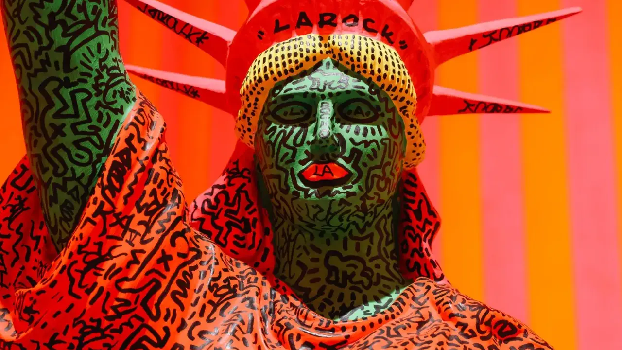 A vibrant Statue of Liberty sculpture, reimagined with Keith Haring's iconic political art style, features a green face with bold black lines and red lips, and an orange robe adorned with his signature figures.