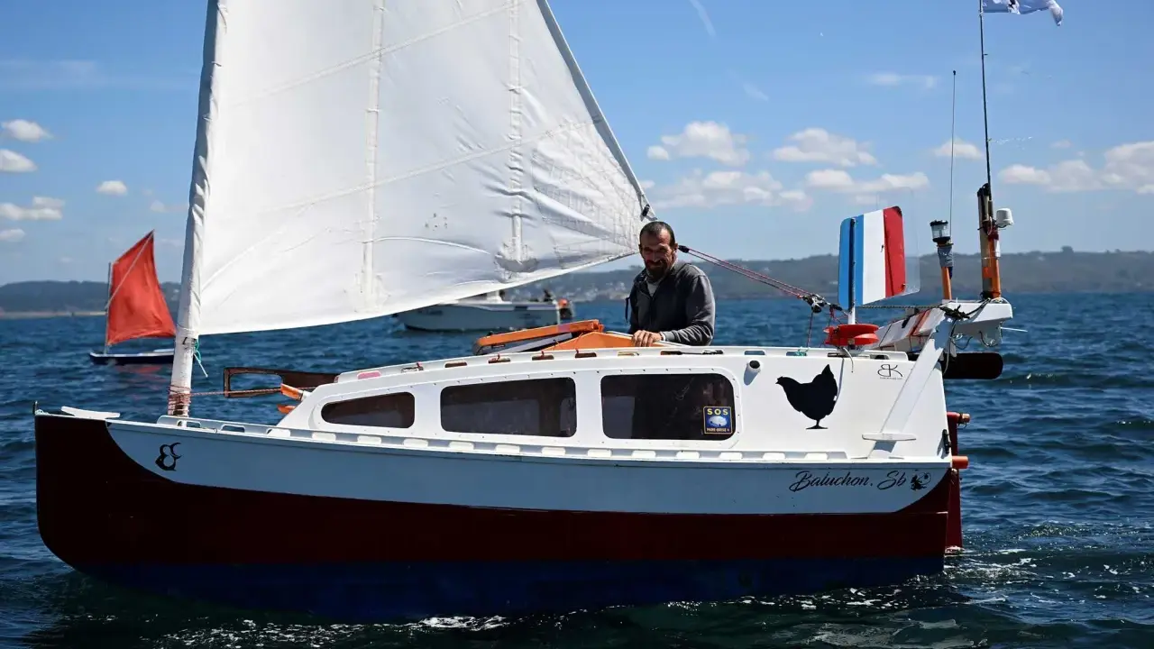 Un homme sourit sur son bateau nommé "Baluchon". Voiles blanches, coque rouge et bleue, drapeau français.