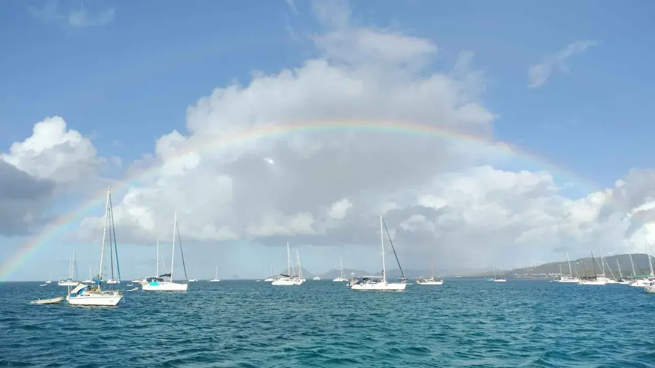 Un arc-en-ciel sur une mer bleue, parsemée de voiliers. L'appel de l'aventure : tout quitter pour vivre sur un bateau.