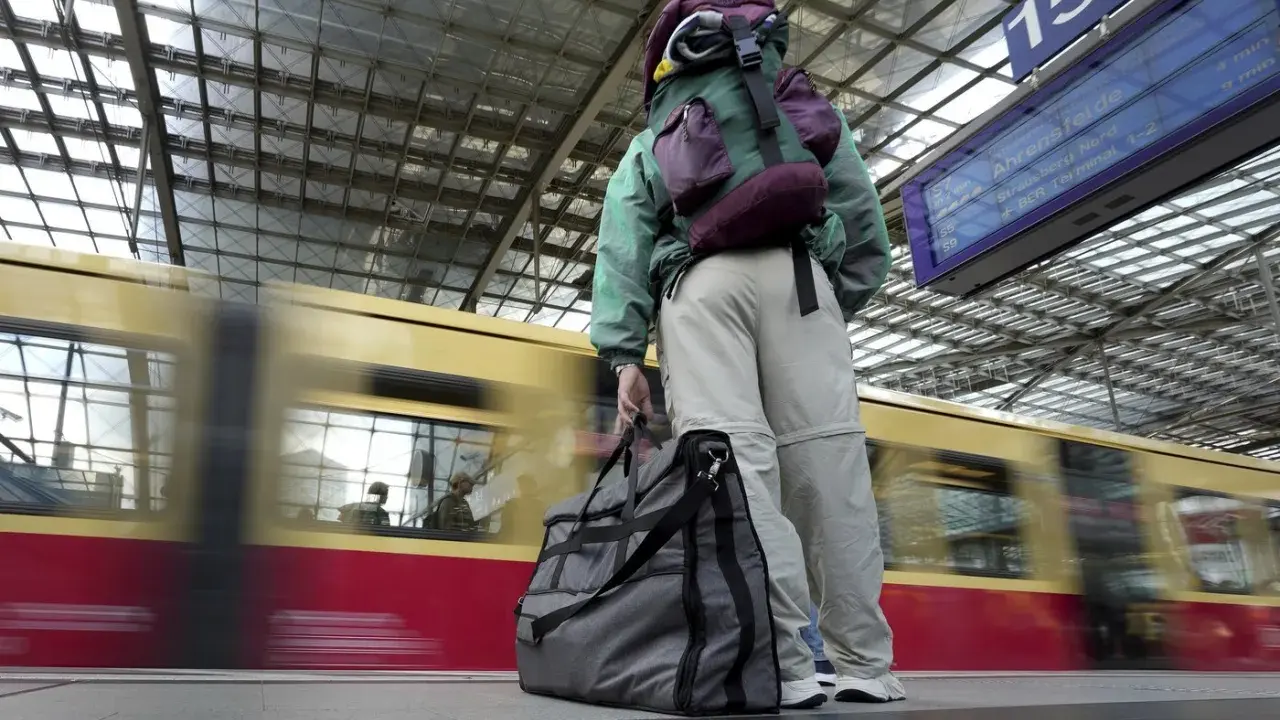 Traveler with backpack and duffel bag waits on a platform as a train speeds by.