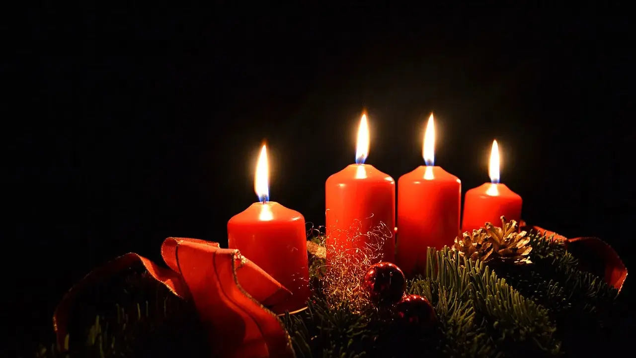 Fyra röda adventsljus brinner i mörkret, en del av en julkrans med granris och röda band. Detta är en visuell representation av vad betyder advent.