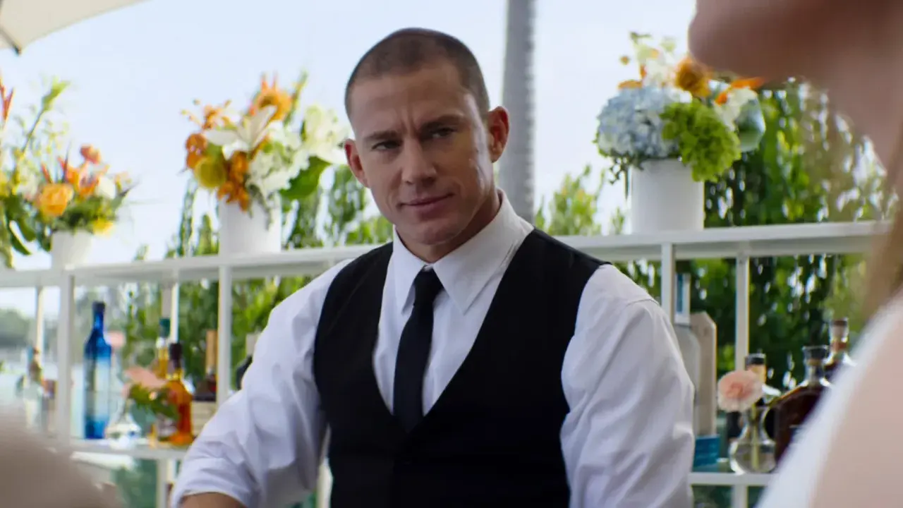 Channing Tatum som bartender i en film. Han ser ut att vara redo att servera drinkar med blommor och flaskor i bakgrunden.