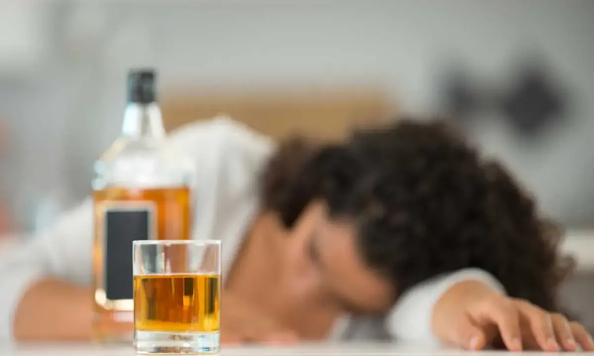 Szklanka z alkoholem i butelka na stole, osoba śpiąca w tle.
