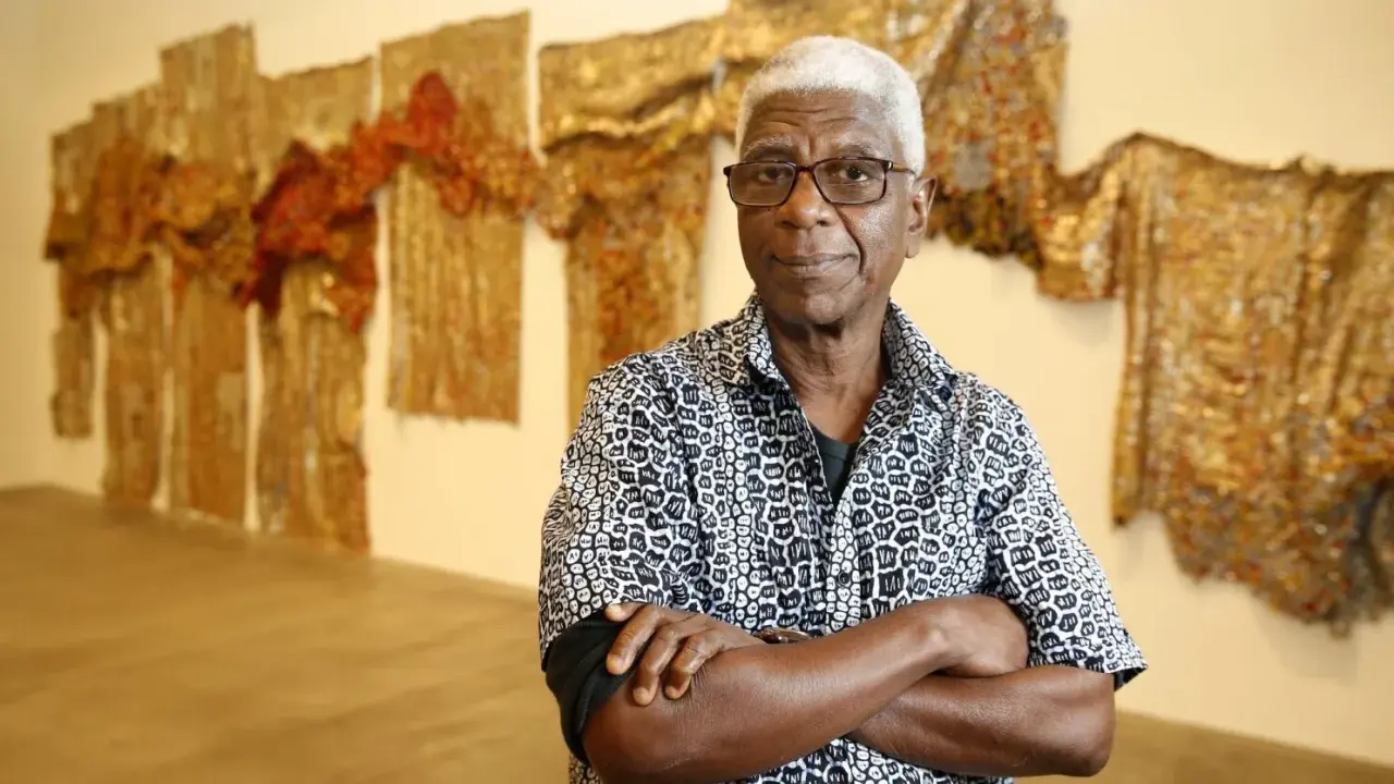 A distinguished man with white hair and glasses, arms crossed, stands before a wall adorned with shimmering, textured artworks, a testament to the enduring legacy of famous artists today.
