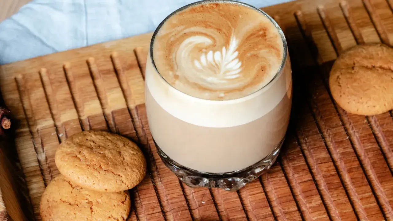 Ein Glas Flat White mit Latte Art neben Keksen auf einem Holztablett. Der Unterschied zwischen Flat White und Cappuccino ist hier deutlich sichtbar.