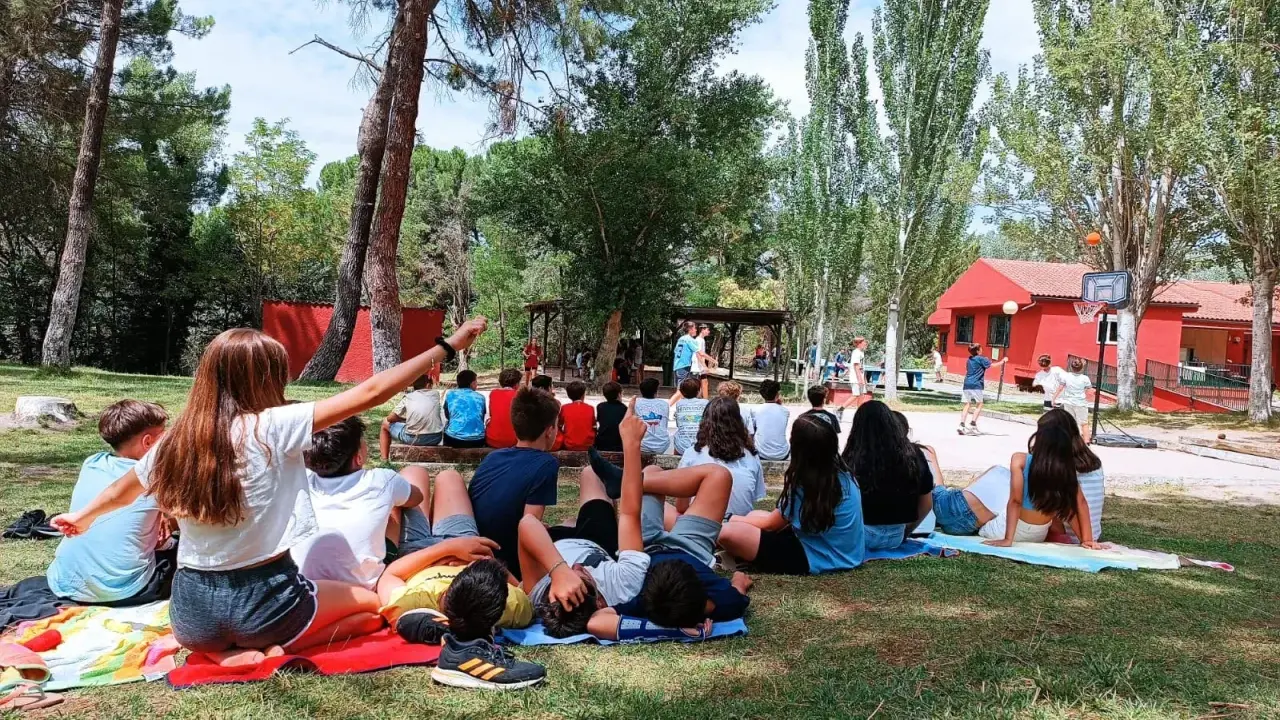 Niños disfrutando de actividades al aire libre en campamentos de verano aula joven. Un grupo observa un partido de baloncesto mientras otros descansan en el césped.