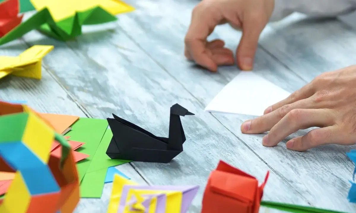 Ręce składają papier, tworząc origami: czarnego łabędzia, czerwoną różę i kolorowe figury. Pokazuje, co można zrobić z papieru.
