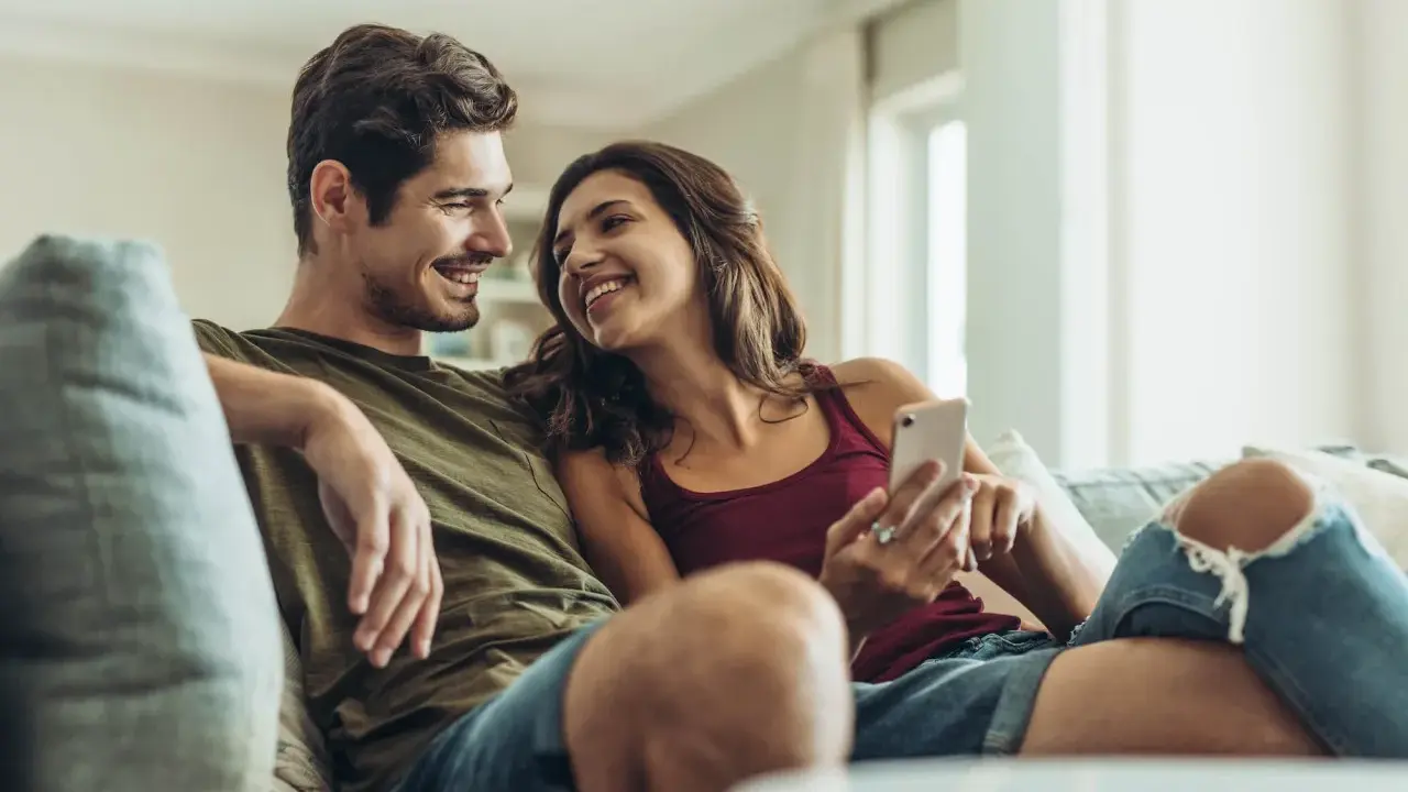 Pareja sonriente en el sofá, compartiendo un momento íntimo. Ella le muestra algo en su móvil, una forma de como expresar sentimientos.