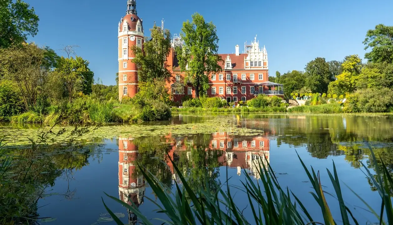 Czerwony pałac z wieżą odbija się w spokojnej tafli wody. Park mużakowski zachwyca zielenią i letnim słońcem.