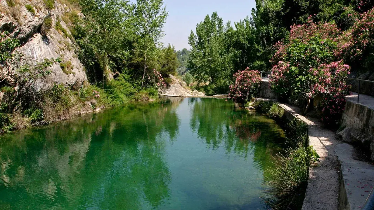 Un remanso de agua verde rodeado de rocas y vegetación exuberante. Un camino serpentea junto al agua, ideal para explorar qué ver en Ontinyent.