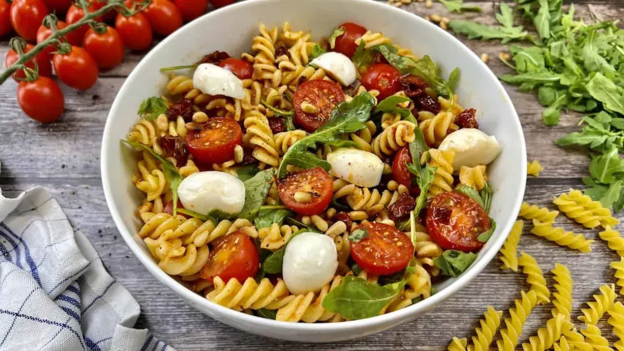 Ein köstlicher Pesto-Salat mit getrockneten Tomaten, Mozzarella-Kugeln und Pinienkernen, serviert in einer weißen Schüssel.