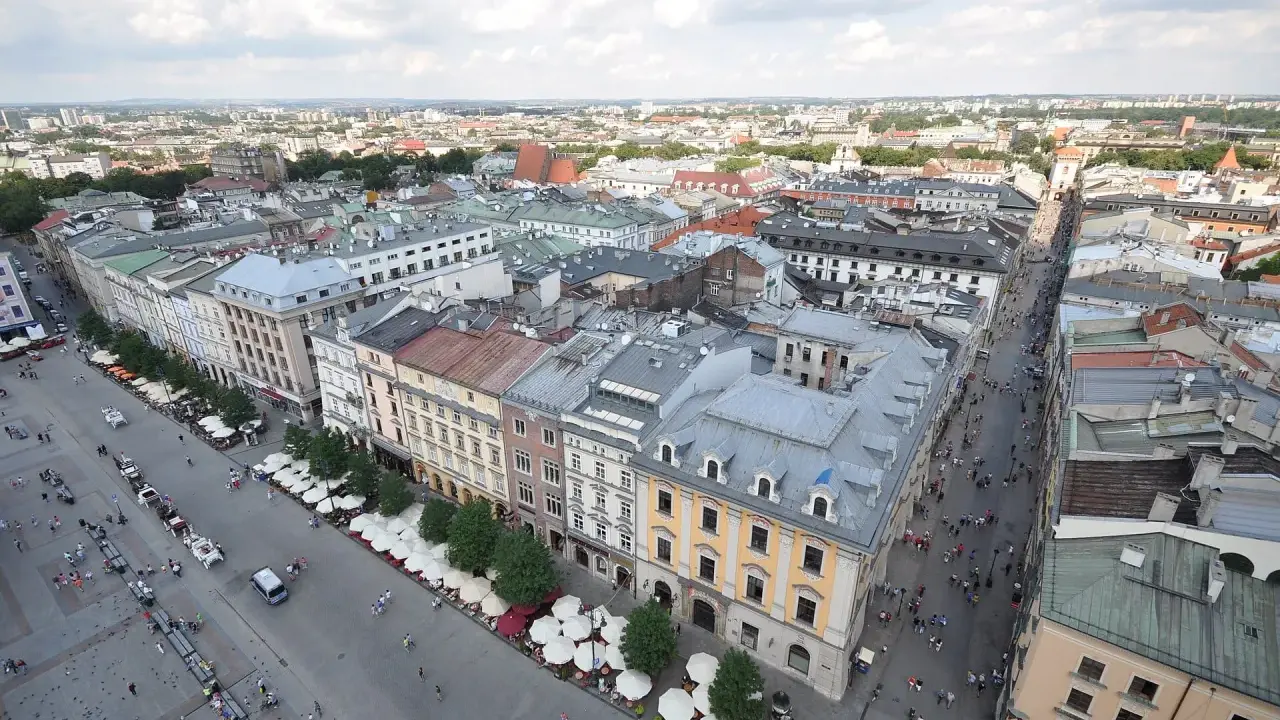 Widok z góry na Rynek Główny w Krakowie. Tłumy ludzi spacerują, a dorożki czekają na turystów. W tle widać zabytkowe kamienice i rozległą panoramę miasta.
