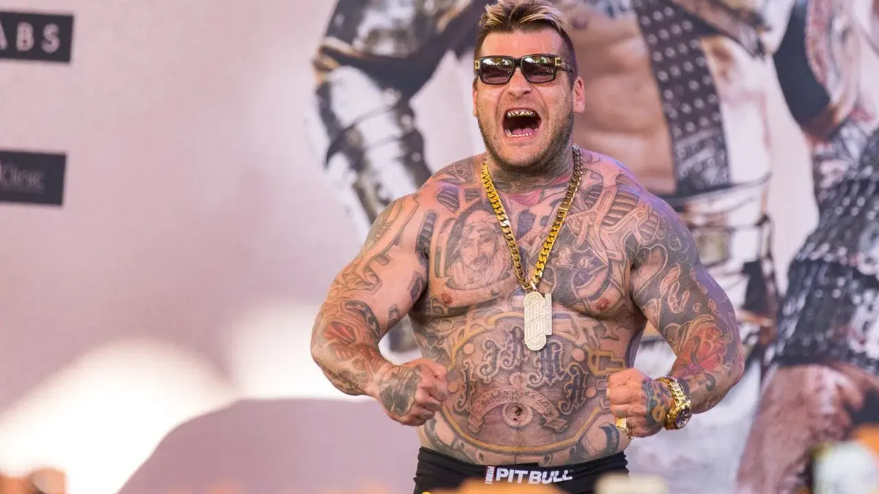 Popek kiedys, wytatuowany mężczyzna w okularach przeciwsłonecznych, z złotym łańcuchem i wisiorem, napina mięśnie.