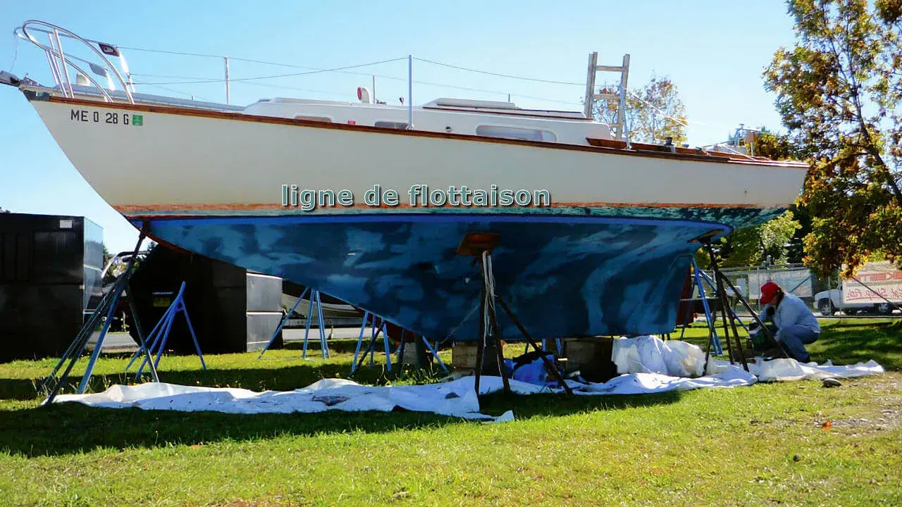 Un bateau est sur des chandelles, prêt pour une peinture. La ligne de flottaison est visible sur la coque bleue. Un homme travaille sur le bateau.