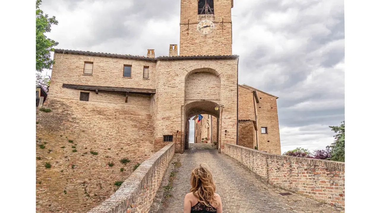 Passeggiata verso il borgo di Montegridolfo cosa vedere: un antico portale in mattoni con torre dell'orologio e un ponte acciottolato.