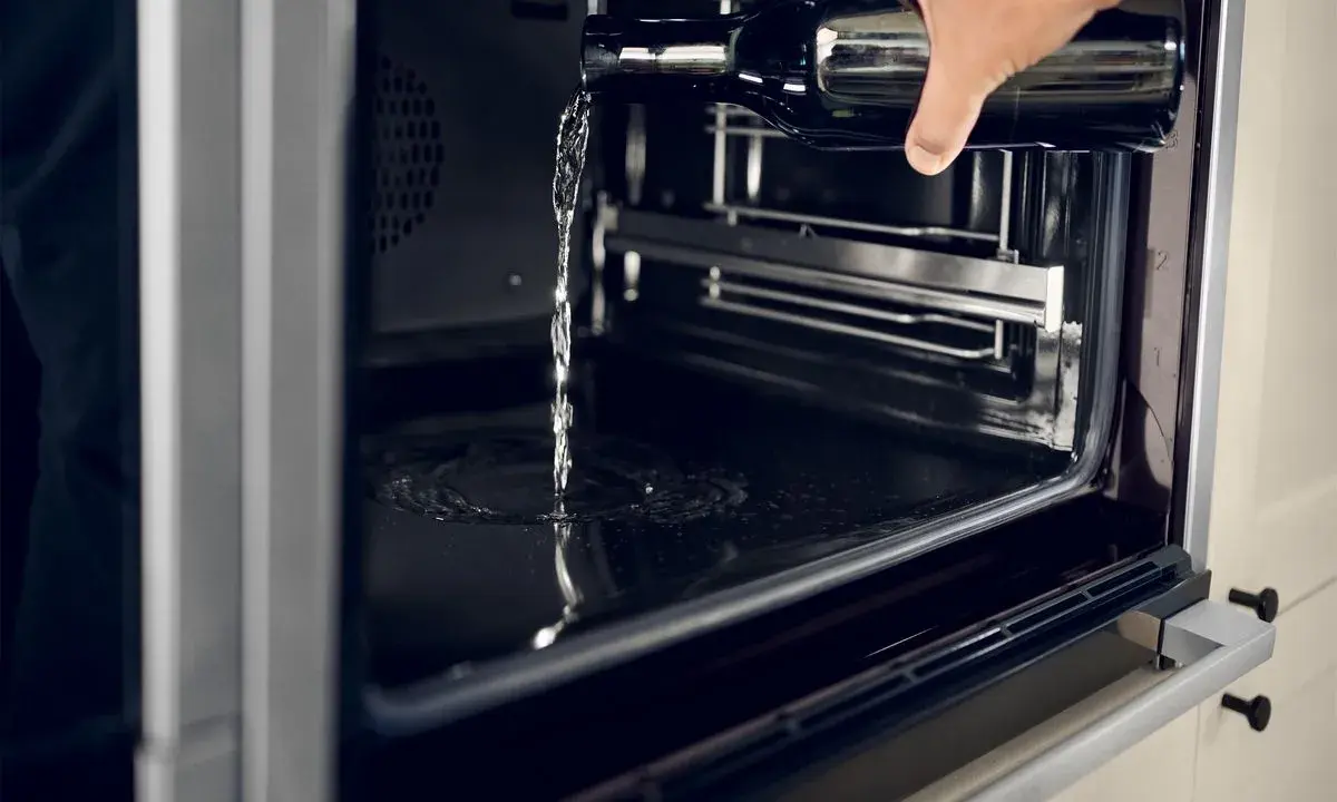 Wasser wird aus einer Flasche in einen modernen **Hydrolyse Backofen** gegossen, um die Reinigung zu erleichtern.