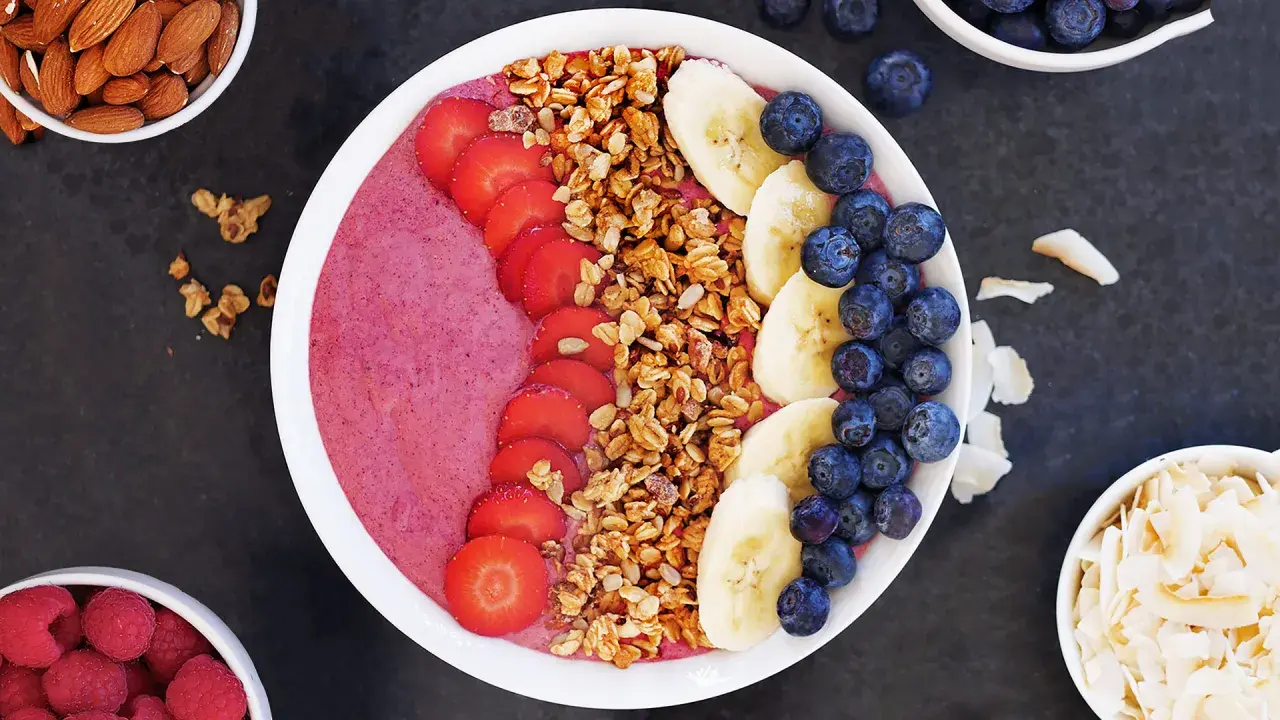 En färgglad acai bowl med jordgubbar, banan, blåbär och granola. Serverad med mandlar och kokosflingor vid sidan.