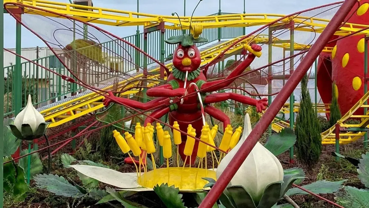 Eine rote Insektenfigur mit vielen Armen steht vor einer gelben Achterbahn.