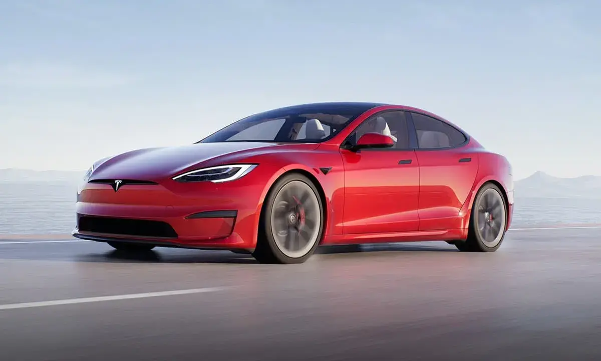 Une Tesla Model S Plaid rouge vif file sur une route côtière, le soleil se reflétant sur sa carrosserie élégante.