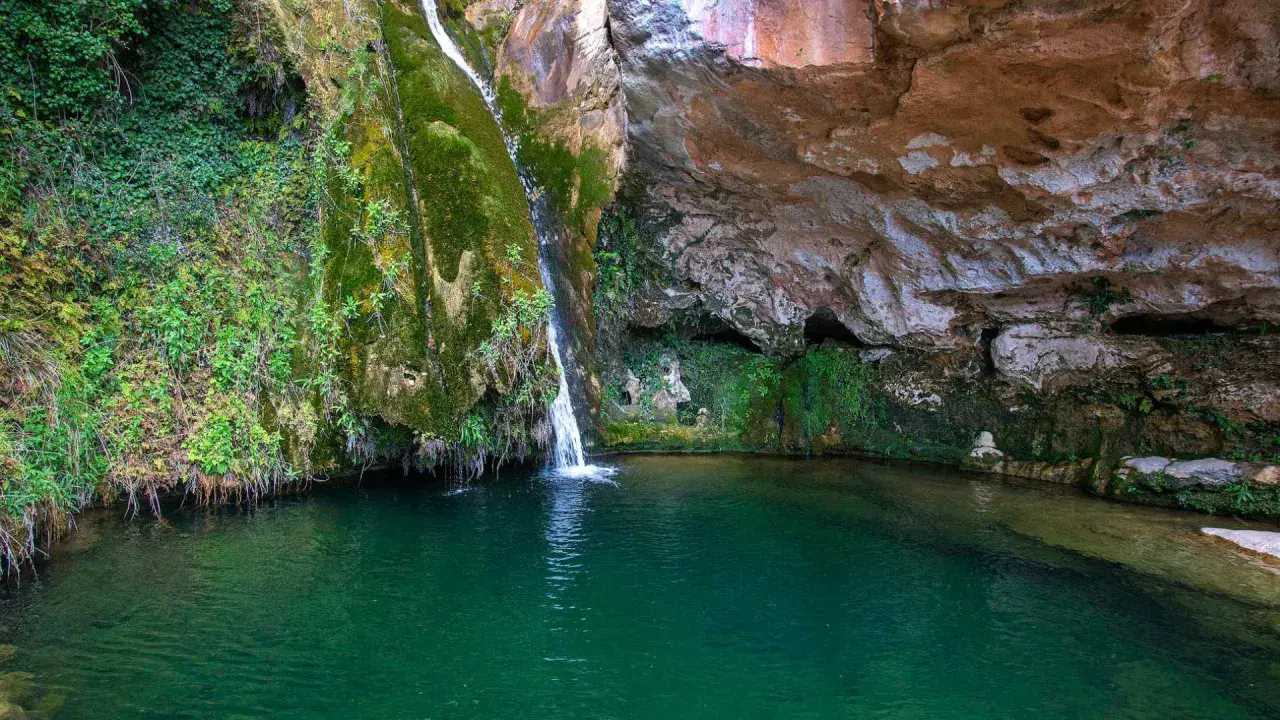 La cascada del río Carbo cae en una poza de agua verde esmeralda, rodeada de rocas cubiertas de musgo y vegetación exuberante.