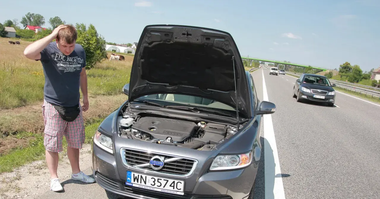 Mężczyzna zmartwiony przy otwartej masce Volvo, prawdopodobnie przegrzany silnik. W tle droga i pola.