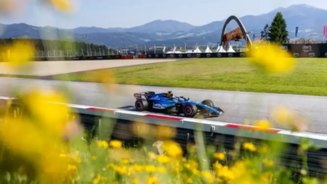 Un coche de F1 azul compite en un circuito soleado, con flores amarillas en primer plano. ¡Un gran resultado f1 hoy!