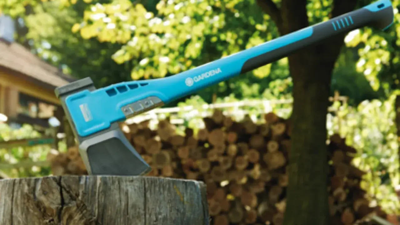 Une hachette bleue et grise est plantée dans un billot, prête à couper du bois. Une pile de bois est visible en arrière-plan.