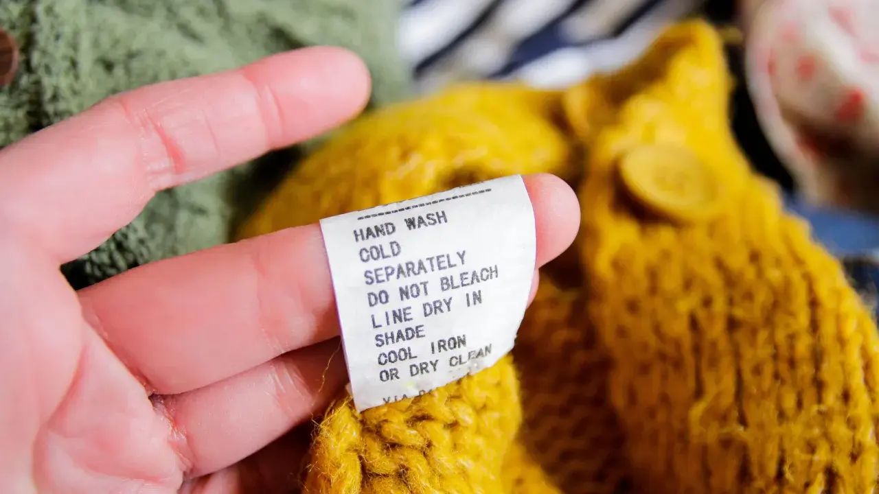 En hand håller en tvättlapp på en gul stickad tröja. Lappen varnar för att ull krymper om den inte sköts rätt.