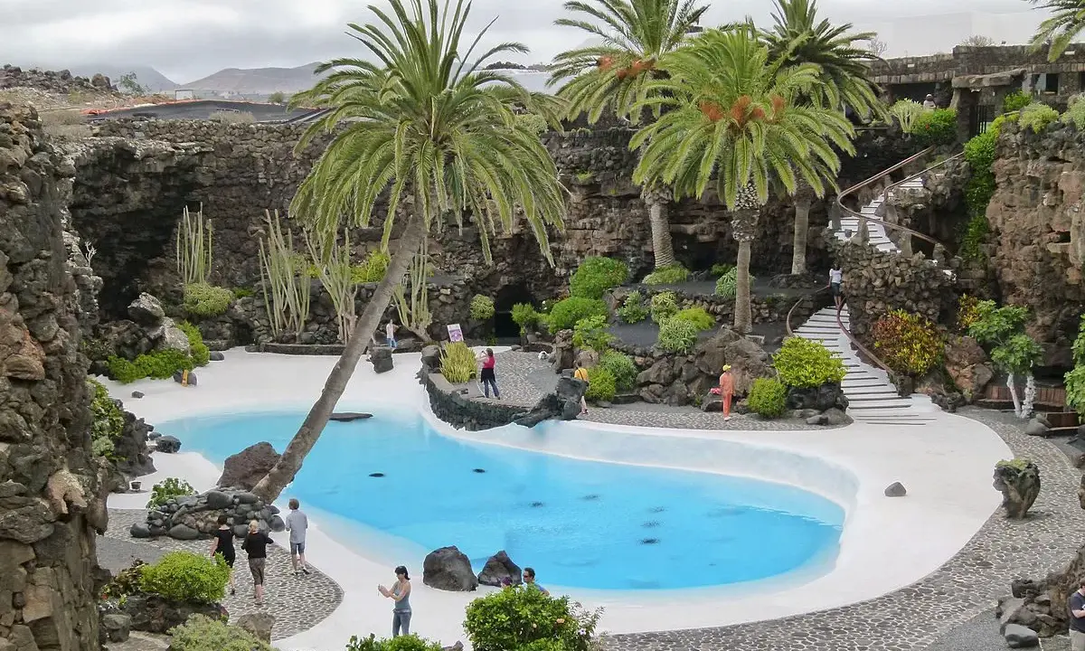 Un oasis de palmeras y cactus rodea una piscina azul en Lanzarote. Ideal para excursiones en Lanzarote con niños, este lugar ofrece un paisaje volcánico único.