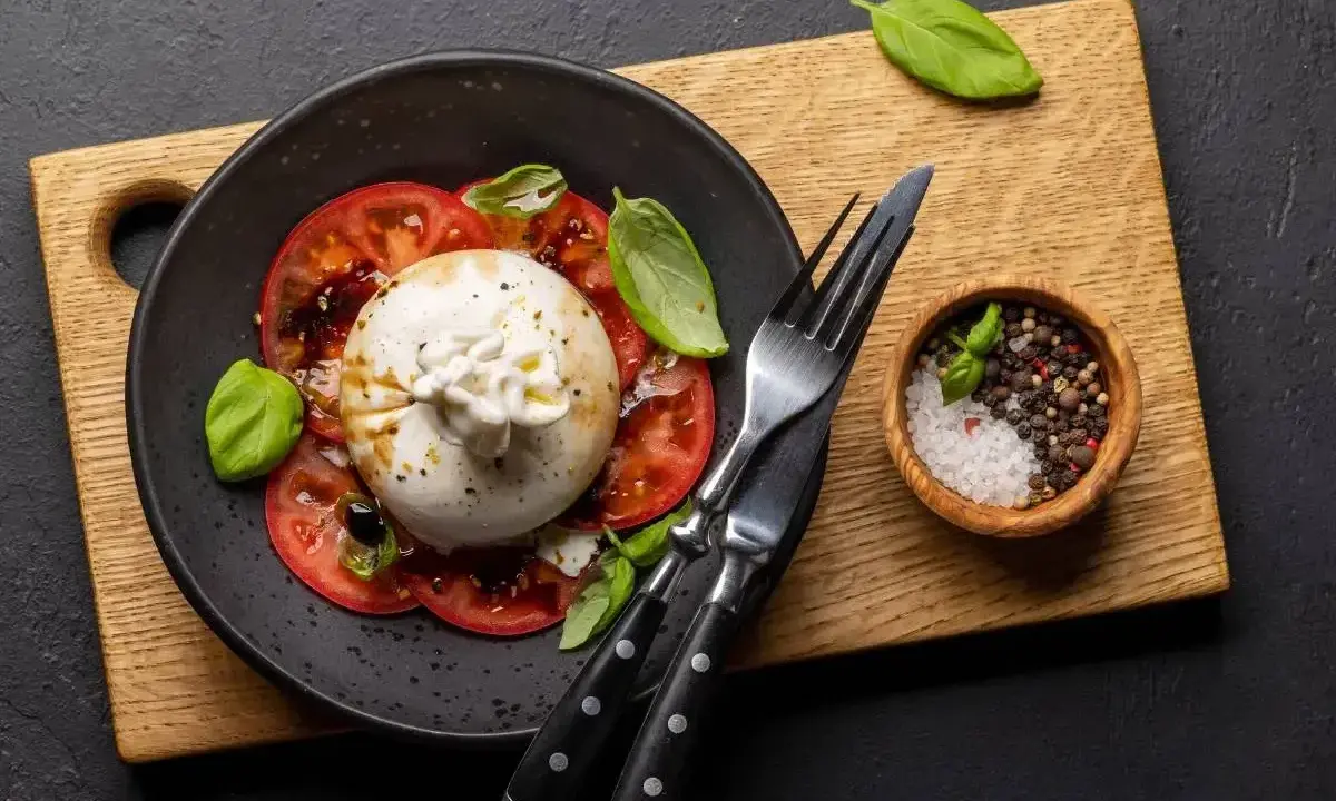 Pyszna burrata z pomidorami, polana oliwą, udekorowana bazylią. Obok miseczka z solą i pieprzem.