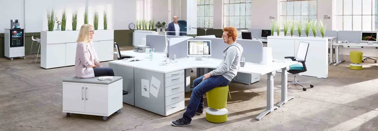 Un espace de travail open space moderne avec des bureaux modulables, des plantes vertes et des sièges colorés. Deux personnes discutent, une troisième travaille sur son ordinateur.