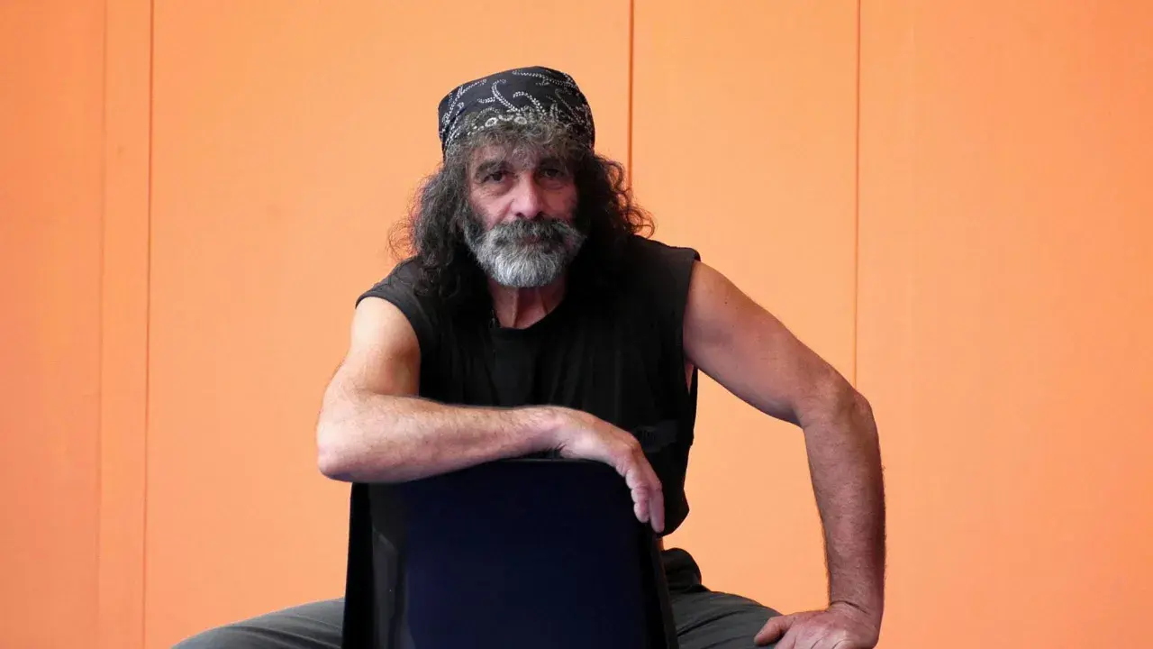 Mauro Corona, con bandana e barba grigia, seduto su una sedia. Immagina i suoi libri in ordine cronologico.