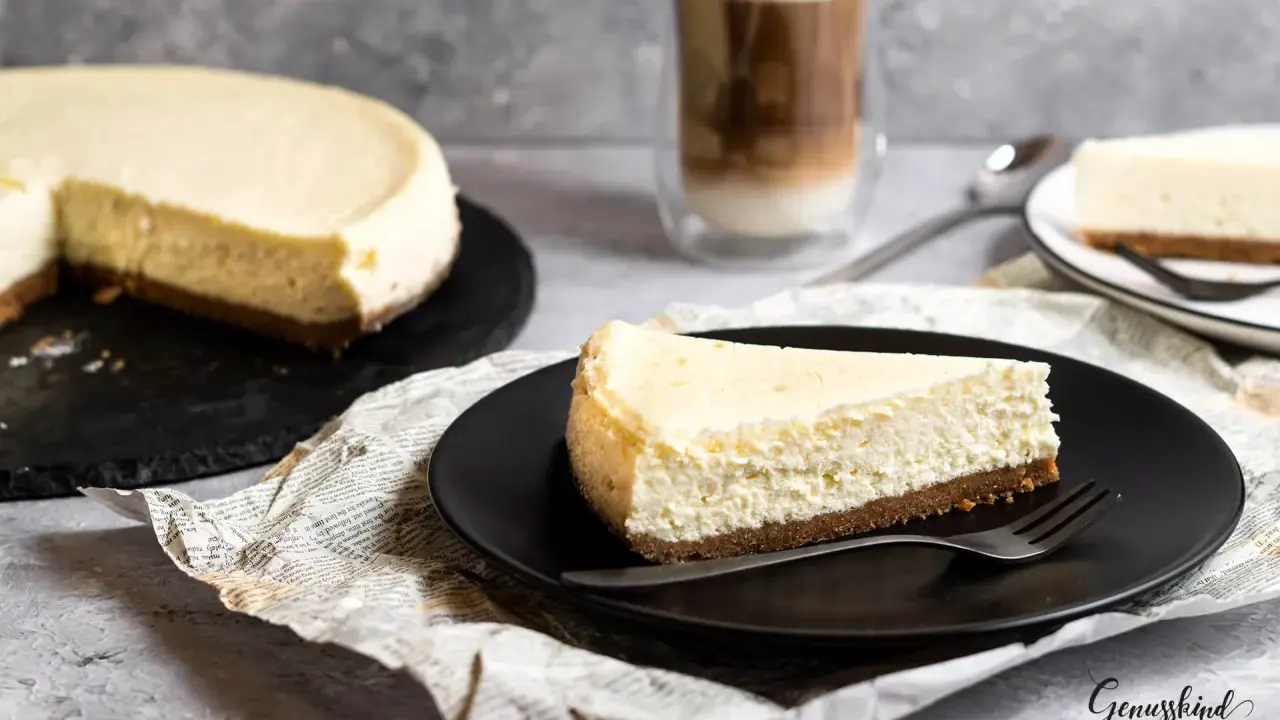 Ein Stück New York Cheesecake auf schwarzer Platte, daneben ein ganzer Kuchen und ein Latte Macchiato.