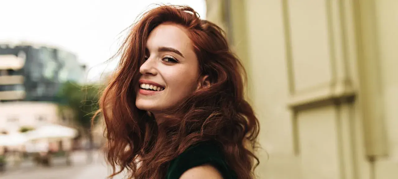 Donna con capelli rossi mossi e un sorriso radioso. La sua chioma è così bella che si può fare la tinta ogni 15 giorni per mantenerla perfetta.
