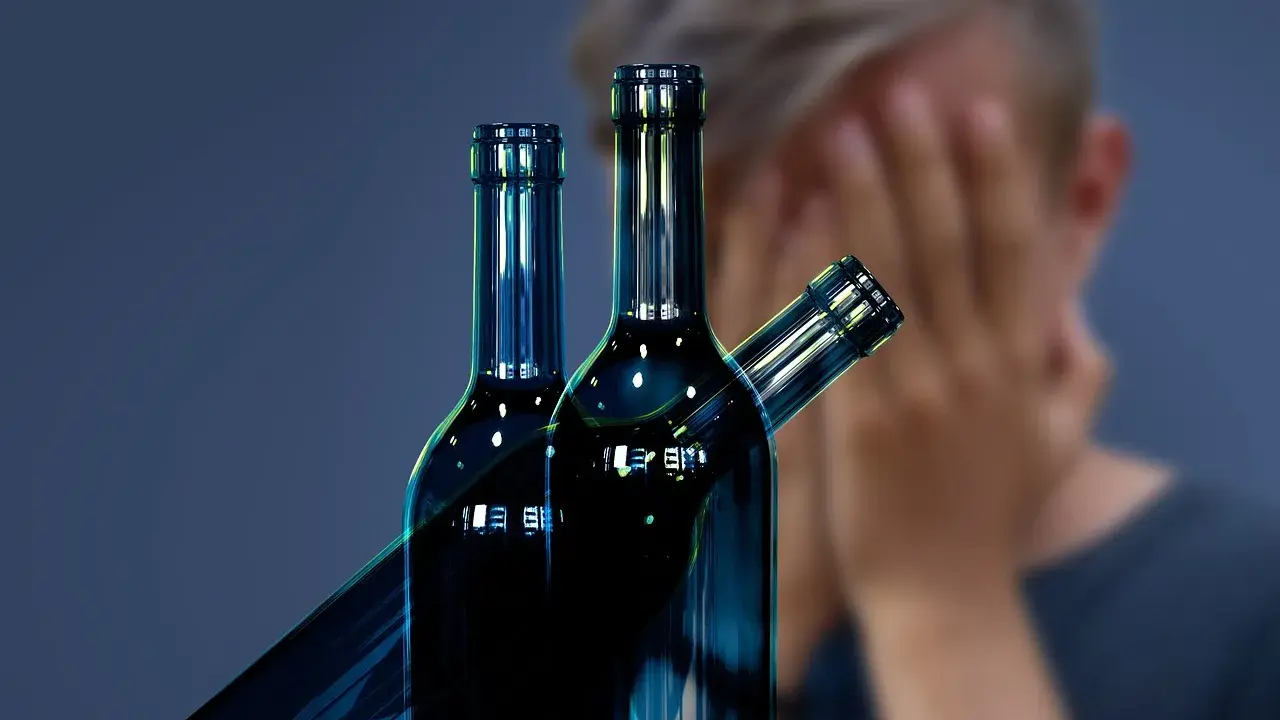 Trzy butelki alkoholu na pierwszym planie, w tle osoba zakrywająca twarz. Symbol walki z nałogiem, nadzieja na odwyk alkoholowy.