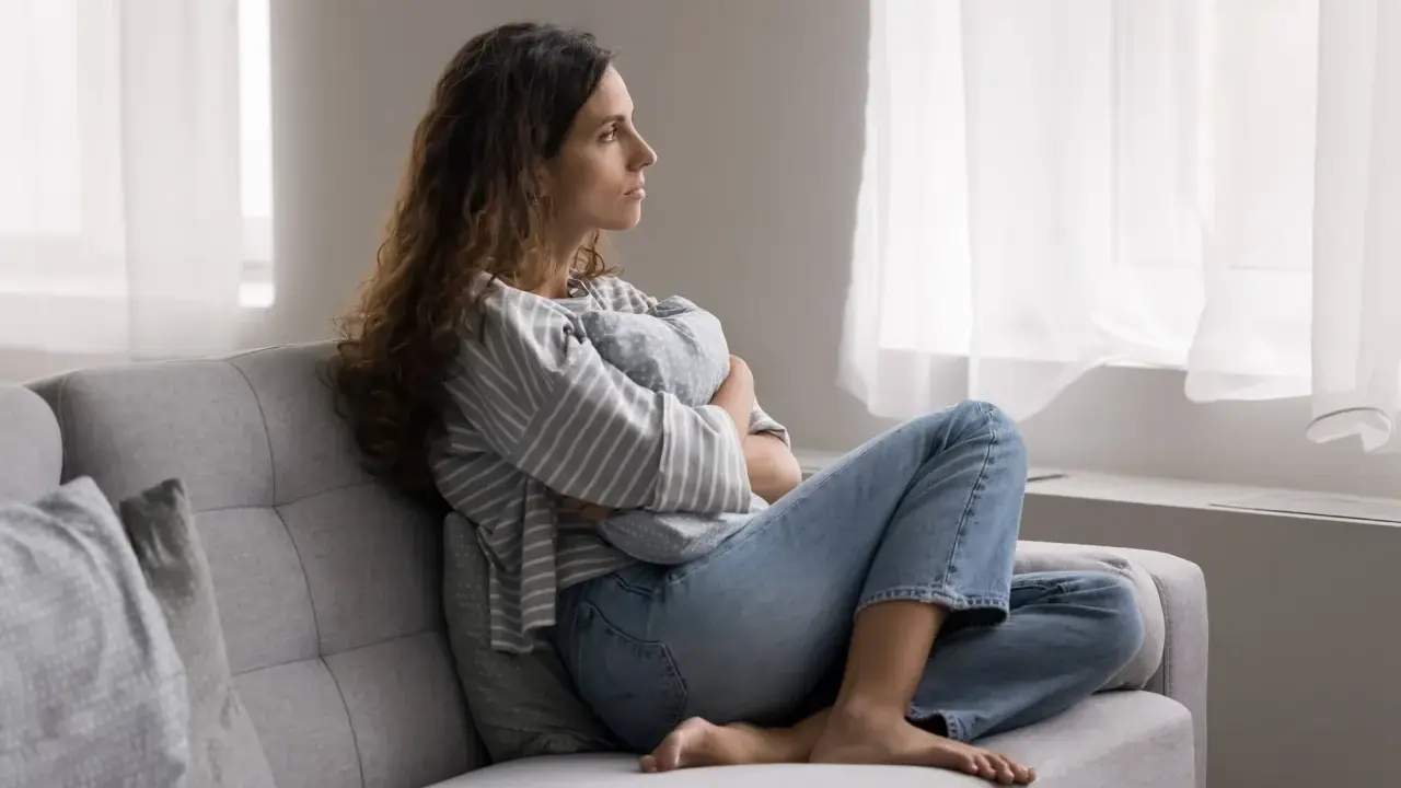 Kobieta siedzi na kanapie, przytulając poduszkę. Zamyślona patrzy przez okno. To pokazuje, po co światu psychoterapia – by odnaleźć spokój.