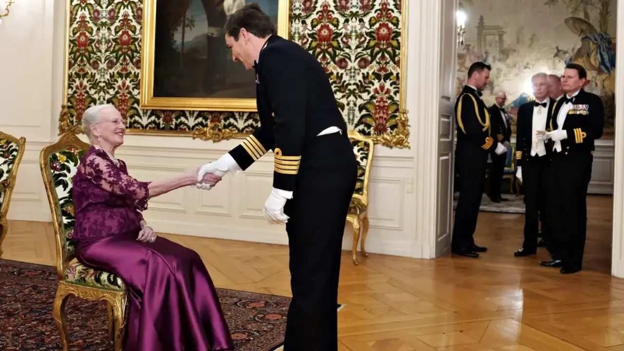 Drottning Margrethe, trots att hon är sjuk, tar emot en officer med ett leende. Hon sitter i en elegant stol, klädd i en lila klänning.