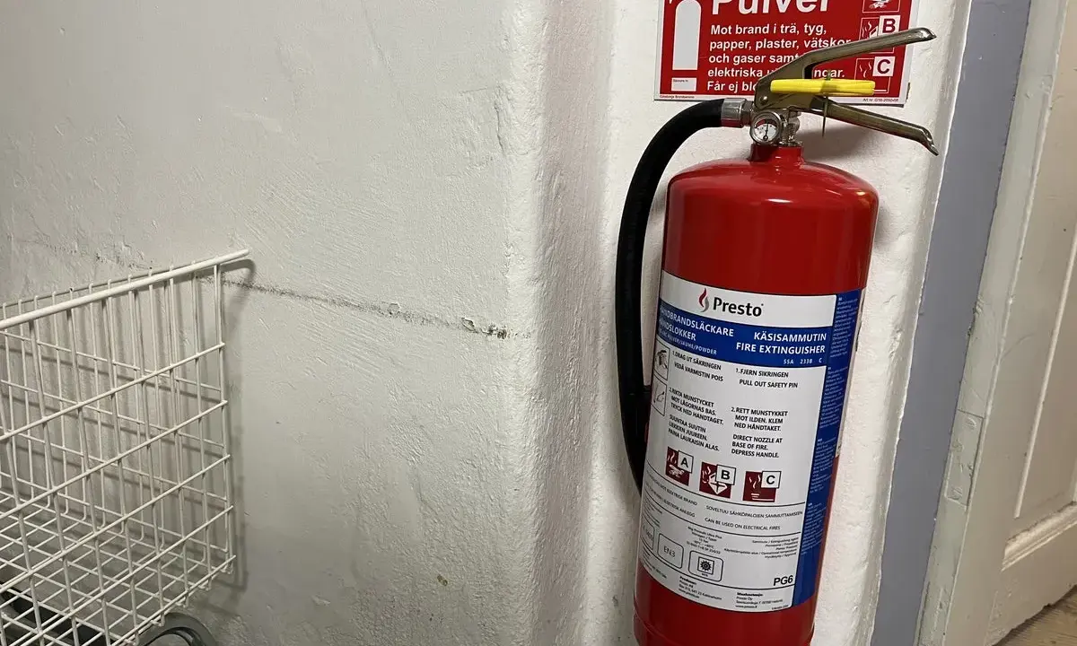Röd brandsläckare hänger på en vit vägg, redo att användas om brandlarm ljuder. Bredvid en vit metallkorg.